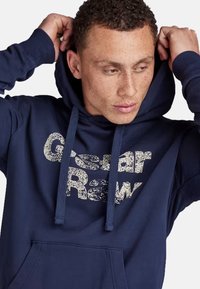Navy blue hoodie with a kangaroo pocket, drawstring hood, and textured "Gstar Raw" graphic on the front. Made from soft fabric.