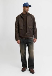 Giacca marrone con zip e colletto, indossata sopra un maglione di lana scura a trama e una camicia a quadri. I jeans neri e le scarpe marroni completano il look.