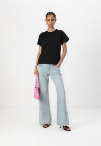 Black cotton t-shirt with a round neck, paired with light blue flared jeans. The model holds a glossy pink handbag and wears black sandals.