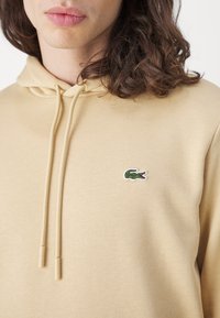 Sudadera beige de tejido suave, con un bolsillo tipo canguro en el frente, cordones y un pequeño logo de cocodrilo verde en el pecho.