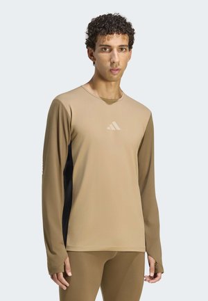 Hombre con camiseta deportiva beige de manga larga con paneles laterales negros y mallas a juego, de pie contra un fondo liso y claro.