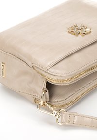 Beige texturovaná umělá kožená taška se zlatými doplňky, s uzávěrem na zip a výrazným dekorativním emblémem se zlatým akcentem.