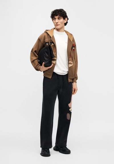 Καφέ φούτερ με κουκούλα και αφηρημένο σχέδιο προσώπου, λευκό μπλουζάκι, μαύρο παντελόνι φόρμας με μπαλώματα και μαύρη τσάντα που κρατιέται κάτω από το μπράτσο.