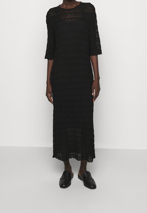 Femme portant une robe midi noire texturée avec des manches aux coudes et des chaussures noires à enfiler, debout devant un fond blanc uni.