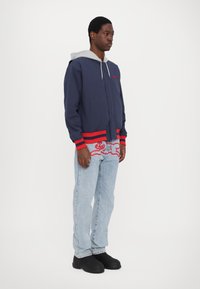 Marineblå jakke med knapper og røde detaljer samt grå hætte, parret med lyseblå jeans med rødt broderet design og sorte sko.