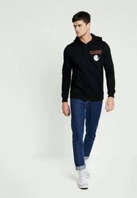 Svart hoodie med en frontficka, prydd med en röd "Popeye"-design. Matchad med blå jeans och vita sneakers, modell promenader på en neutral bakgrund.