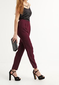 Pantalon bordeaux sur mesure avec plis, associé à un haut en satin noir et des talons compensés noirs. Pochette avec un motif géométrique.