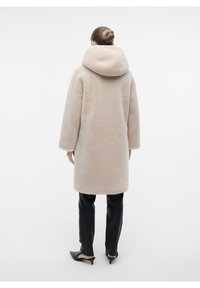 Cappotto teddy beige chiaro con un ampio cappuccio, tessuto testurizzato, maniche lunghe e taglio dritto. Indossato sopra pantaloni neri e scarpe.