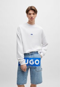Sweatshirt blanc oversized avec un petit logo bleu sur la poitrine, associé à un short en denim bleu arborant un grand graphisme bleu "HUGO".