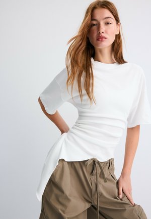 Jeune femme aux longs cheveux roux porte un haut blanc ample et un pantalon cargo kaki, posant avec une main sur la hanche devant un fond uni.