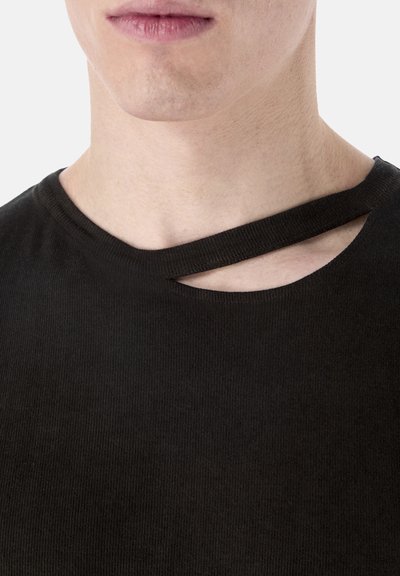 Jeune personne portant un haut noir à côtes avec une découpe asymétrique au niveau de la clavicule.