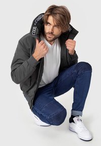 Grey bomber jacket with ribbed cuffs, white T-shirt, dark blue jeans with ribbed detailing, and white sneakers. Model kneeling, facing slightly left.