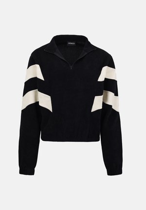 Černý svetr s polovičním zipem, s bílými cikcak pruhy na rukávech a žebrovanou texturou, zobrazený na neutrálním pozadí.