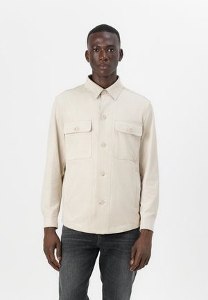 Beige button-up shirt jacket made of lightweight fabric, featuring two chest pockets and a collared neckline. Smooth texture, straight cut.