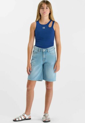 Fille debout portant un haut bleu sans manches avec découpe, un short en jean clair jusqu'aux genoux et des sandales argentées sur fond blanc.