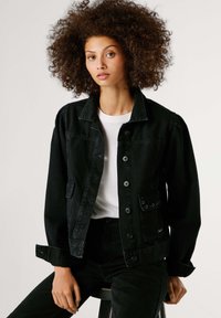 Crna traper jakna s ovratnikom, prednjim kopčanjem na gumbe, džepovima i zavrnutim manšetama. Stilizirana s bijelom majicom ispod.