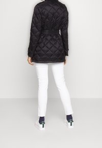 Quiltad svart jacka med bälte, som har krage och mönstrad textur. Kombineras med vita byxor och vita sneakers med gröna detaljer.