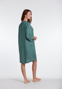 Losse, knie-lange jurk in diep teal groen, gemaakt van gladde stof met lange mouwen en zijzakken. Minimalistisch ontwerp.