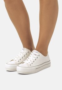 Zapatillas de lona blancas con un perfil bajo, punta redonda y suela de goma texturizada. Presentan ojales metálicos y rayas decorativas grises.
