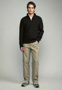 Schwarzer Rippstrickpullover mit Reißverschlusskragen, beigefarbene Hose und graue Sneakers. Das Modell steht aufrecht mit entspannten Händen an den Seiten, neutraler Hintergrund.