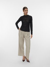 Nő áll, keze bézs széles szárú nadrág zsebében, fekete virágmintás csipke hosszú ujjú blúzt viselve, sötét hegyes sarkú cipővel.