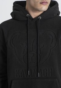 Czarna polarowa bluza z kapturem z teksturowanym logo niedźwiedzia i wyembossowanym napisem "KAVANAGH". Ma sznurki z metalowymi końcówkami oraz kieszeń typu kangur.