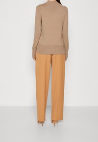 Beige Strickpullover mit geripptem Saum, kombiniert mit hochgeschnittenen beige Hosen. Glatte Textur, körperbetontes Design und dezente Rückendetails.