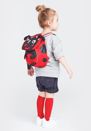 Red and black ladybug-themed backpack with polka dots, plush antennae, and a smiling face, worn by a child in grey top and navy shorts.