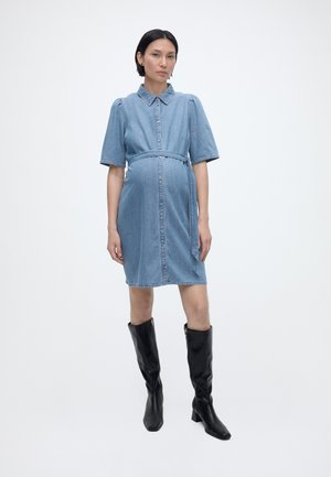 Έγκυος γυναίκα που στέκεται, φοράει γαλάζιο τζιν φόρεμα με ζώνη και κοντά μανίκια, καθώς και μαύρες μπότες μέχρι το γόνατο, σε απλό λευκό φόντο.