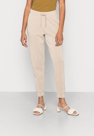 Kvinde iført beige joggerbukser med snøre, kombineret med hvide åbne blokhælssandaler og en brun top, stående mod en ensfarvet baggrund.