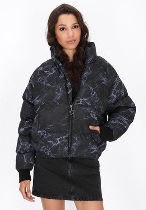 Schwarze gepolsterte Jacke mit einem marmorierten Muster in Blau. Hat einen hohen Kragen, einen Reißverschluss und gerippte Bündchen. Getragen über einem schwarzen Jeansrock.