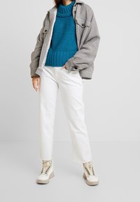 Veste à carreaux surdimensionnée, pull en maille bleu texturé, pantalon blanc à jambes droites et bottes beiges à lacets avec semelle épaisse.