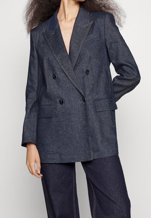 Vrouw draagt een donkerblauw dubbelbreasted blazer met zwarte knopen en bijpassende wijde broekspijpen tegen een effen achtergrond.