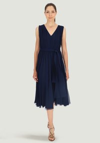 Granatowa sukienka bez rękawów z dekoltem w serek, plisowanym wzorem i detalem w postaci paska. Posiada zwiewną spódnicę z przezroczystym wykończeniem i sandały na rzemykach.