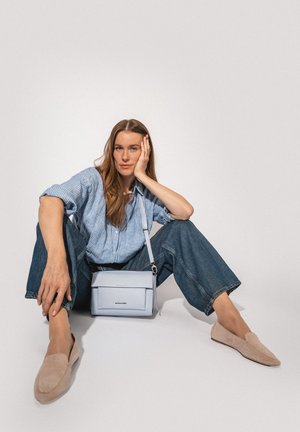 Frau sitzt auf dem Boden in blau gestreiftem Hemd, weit geschnittenen Jeans, beigen Loafers, mit einer hellblauen Umhängetasche, die auf dem Schoß liegt, vor einfarbigem Hintergrund.