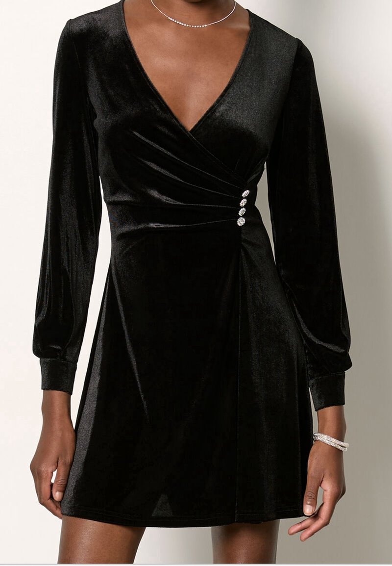 Robe portefeuille longue en velours noir à manches longues avec trois boutons décoratifs en strass à la taille, portée avec un collier délicat et un bracelet.