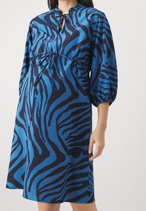 Kvinde iført blå og sort abstrakt zebra-print kjole med trekvart puffede ærmer og snorebind ved halsudskæring og talje.