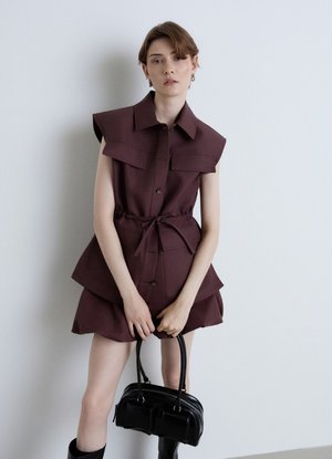 Giovane donna con vestito bordeaux senza maniche che tiene una borsa nera, in piedi davanti a un muro grigio chiaro semplice.