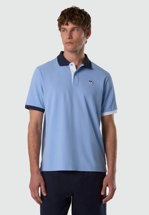 Uomo che indossa una polo azzurro chiaro con colletto e polsini blu navy, in piedi su uno sfondo chiaro semplice, guarda di lato.