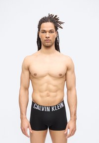 Μυώδης άντρας με ντραντλόκς που φοράει μαύρα εσώρουχα Calvin Klein boxer, στέκεται μπροστά σε απλό φωτεινό φόντο.