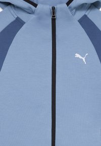 Svijetloplavi dukserica s crnim patentnim zatvaračem i detaljima, s raglan rukavima u tamnijoj nijansi plave. Bijeli logo u središtu na prsima.