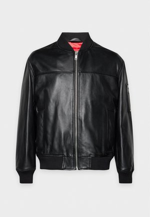 Un homme porte une veste bomber noire en cuir, un T-shirt blanc, un jean foncé et des bottes noires sur un fond clair et uni.