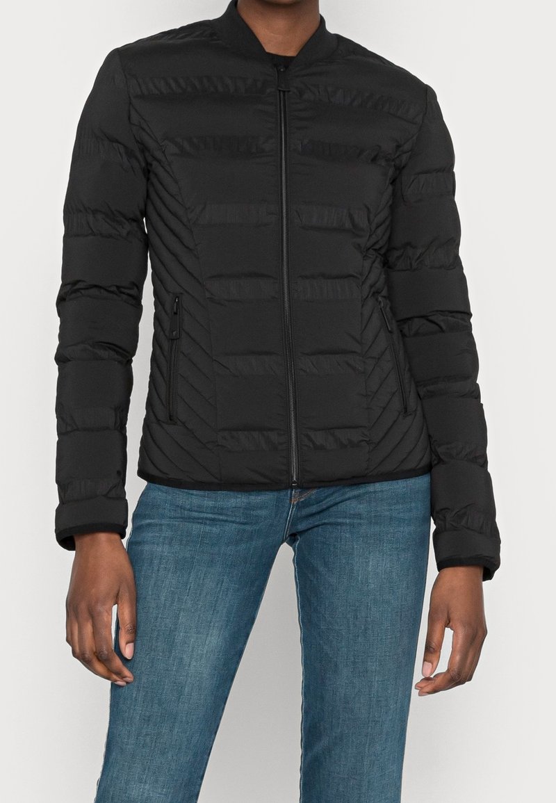 Personne portant une veste noire matelassée zippée avec des poches latérales zippées et un jean bleu, debout devant un fond clair uni.