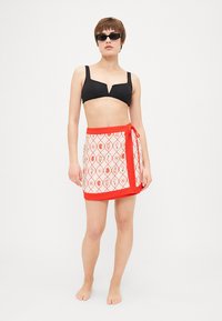 Svart bikini topp med V-hals design, kombinert med en hvit skjørt med rød blomsterdesign og rød knyteliv. Glatt stofftekstur.