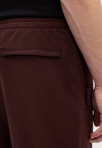 Bordo joggeri ar elastīgu jostu, ar aizmugurējo kabatu un gludu tekstūru. Sānskats parāda tuvplānu uz audumu.