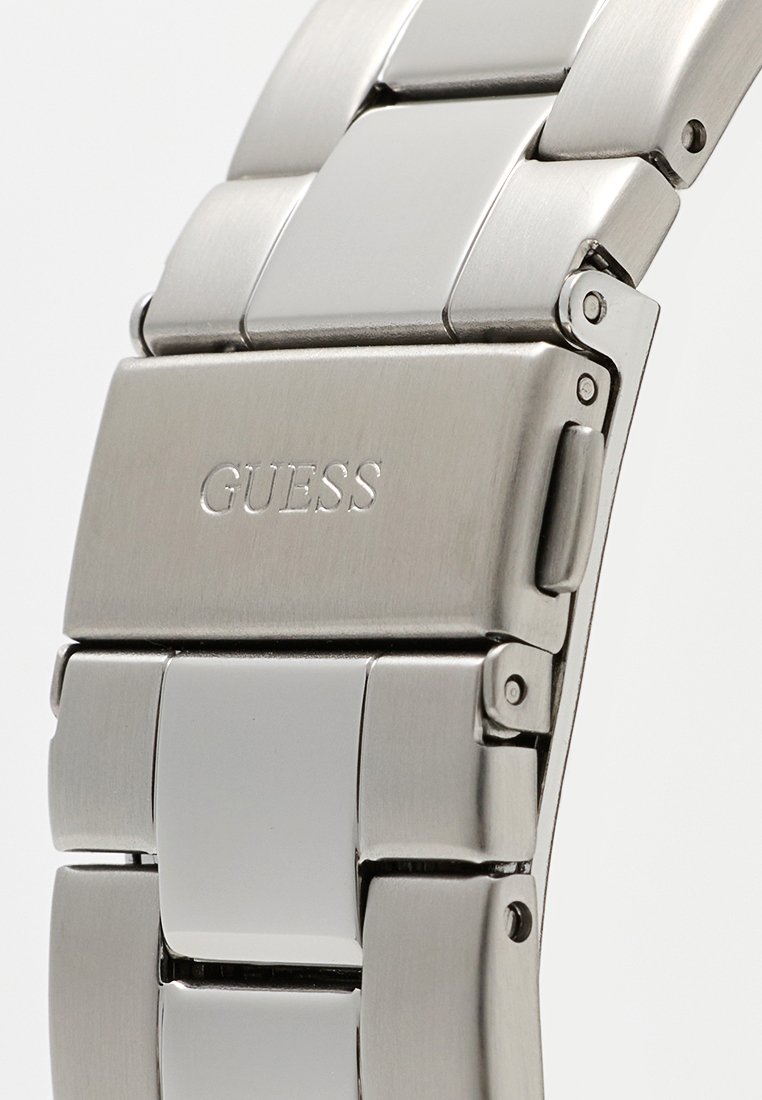 Reloj de acero inoxidable con acabado plateado, con eslabones entrelazados, superficie pulida y un cierre grabado con la palabra "GUESS".