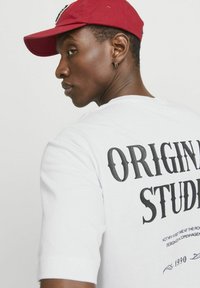 Camiseta blanca con "ESTUDIANTE ORIGINAL" impreso en letras negras en negrita en la parte posterior. El modelo lleva una gorra roja. Manga corta, material de algodón.