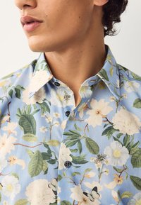 Lyseblå skjorte med blomstermønster bestående af store hvide og gule blomster, grønne blade samt små bier og sommerfugle. Kort krave, knapper ned foran.