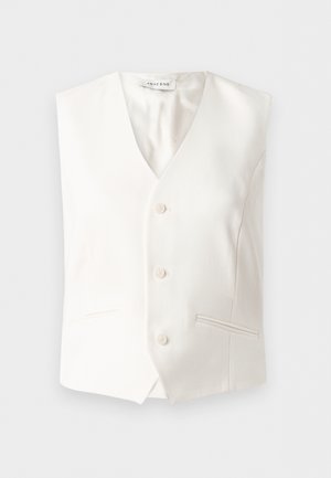 Nő fehér ujjatlan V-nyakú mellényben és bőszárú nadrágban, fekete bőrtáskát tart, fekete magassarkú szandállal párosítva.