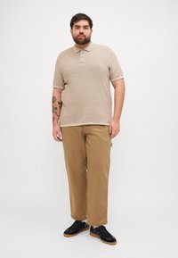 Polo en tricot beige avec un col, patte de boutonnage à trois boutons, associé à un chino kaki et des baskets noires. Tatouages visibles sur le bras gauche.